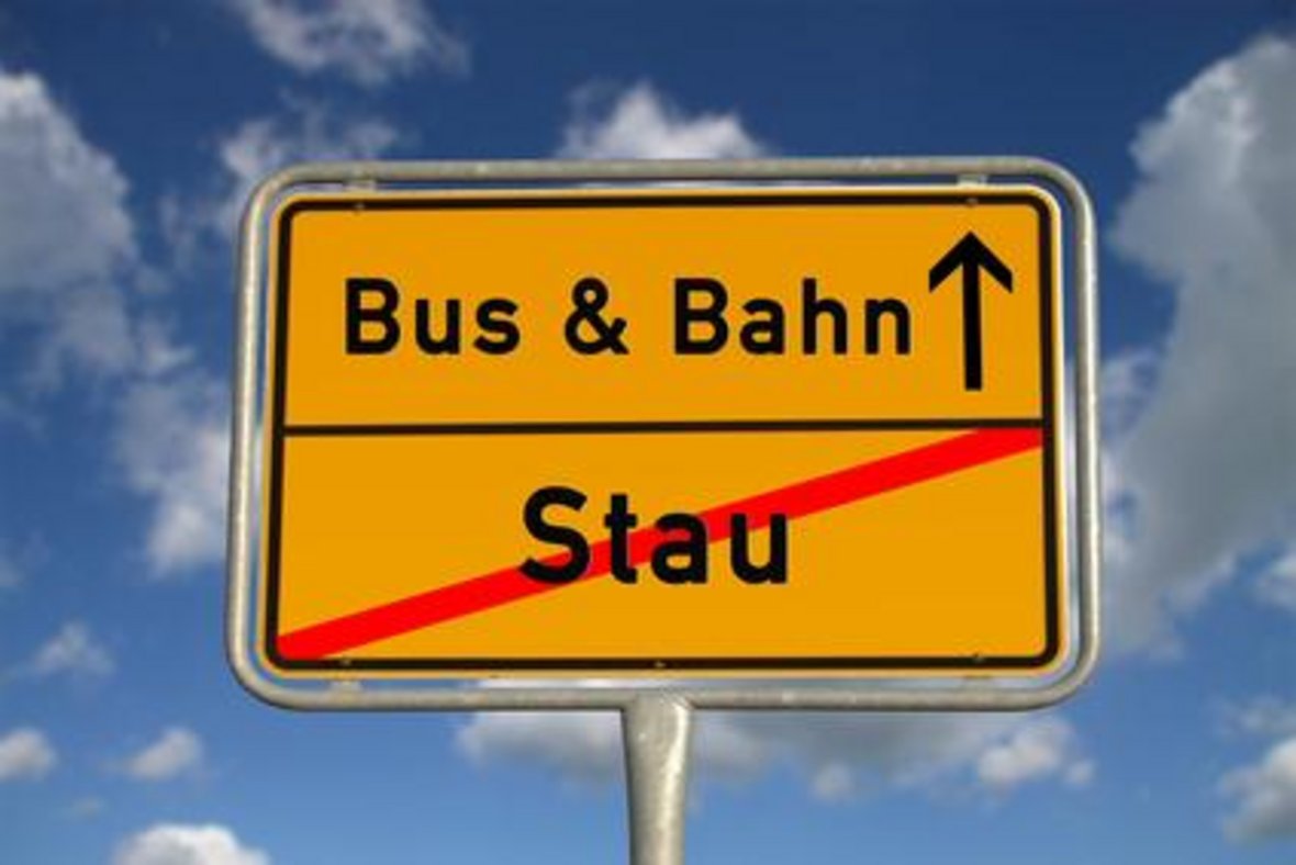 Ein gelbes Ortsschild mit der Aufschrift "Bus & Bahn" und einem Pfeil nach oben, der für die Weiterfahrt steht. Darunter steht das Wort "Stau", das mit einem roten Querbalken durchgestrichen ist. Das Schild ist vor einem blauen Himmel mit einigen Wolken zu sehen. 